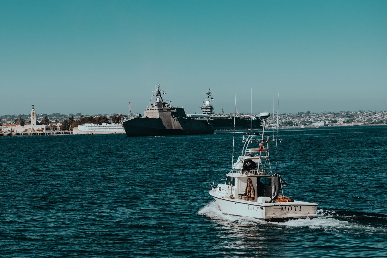 A motorboat navigates the sea with naval ships anchored near a coastal city under a clear sky.