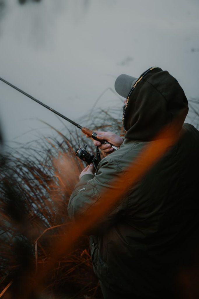A man in a hoodie fishes by a calm lake surrounded by autumn foliage.