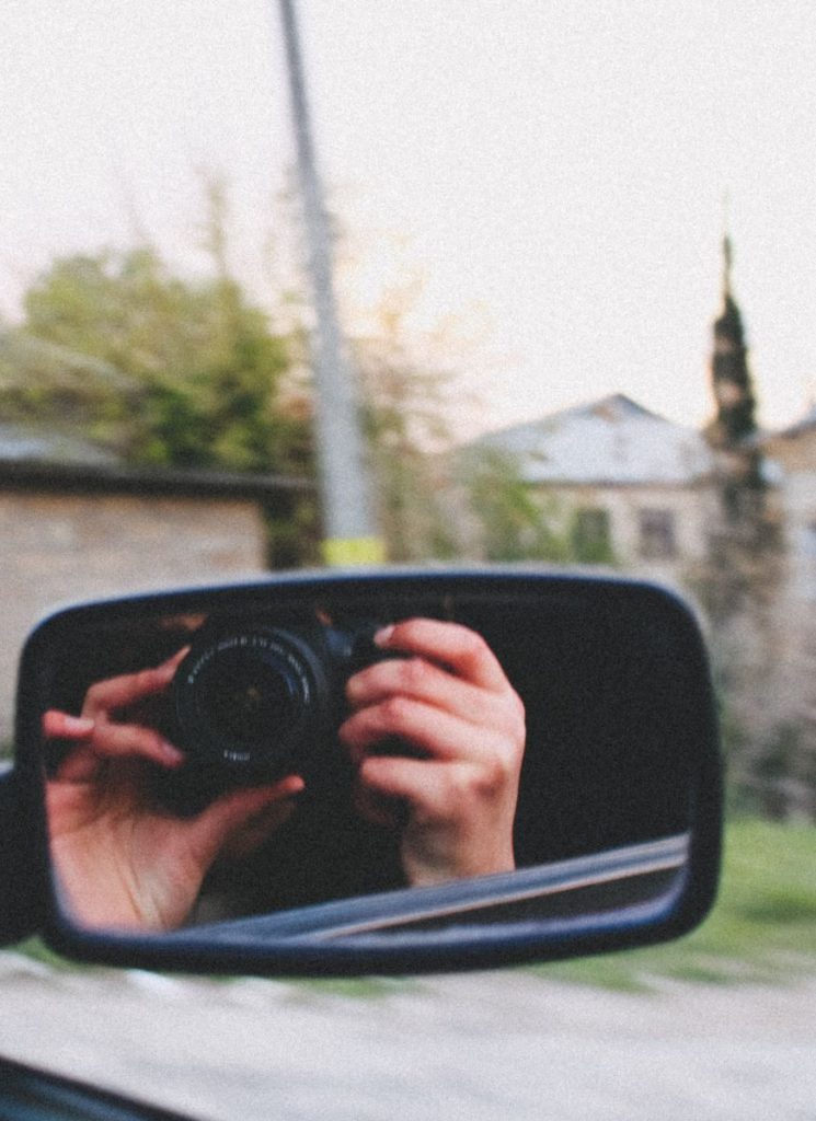 Mirror reflection of a photographer capturing rural scenery through car mirror.