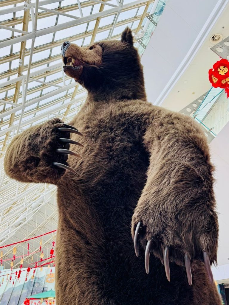 A large bear statue displayed indoors, surrounded by modern architecture and festive decor.